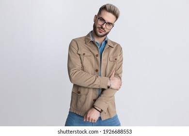 Cool Unshaved Fashion Guy In Jacket Wearing Eyeglasses, Touching Elbow And Holding Arm In A Fashion Pose Against Gray Background In Studio, Portrait