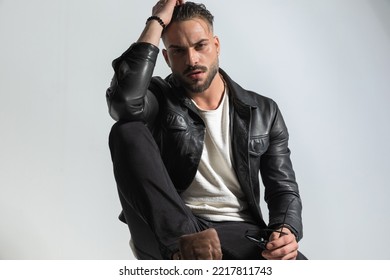 Cool Unshaved Man With Wet Hair Holding Elbow On Knee And Posing In Front Of Grey Background In Studio 