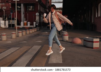Cool woman in beige jacket jumping at street. Girl with short hair in jeans, glasses and light sneakers with backpack and cup of tea posing outside..