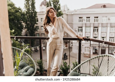 Cool woman in beige outfit smiling on terrace. Happy young lady in stylish pants and shirt poses on balcony and enjoys sunny weather
