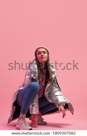 A cool woman in a shiny silver down jacket and a knitted hat squatted down, Studio shot on a pink background, copy space.
