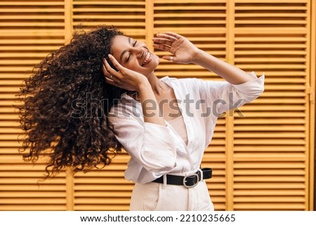Cool young african girl waving her hair closed eyes smiling with teeth on yellow background. Model brunette curly hair wears singlet, shirt and white jeans. Concept enjoyable mood.