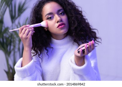 Cool young beautiful mixed-race girl applying makeup with brush in neon light in make-up room. Curly 20s biracial female do natural modern make up, put cosmetics powder on face. Daily beauty routine. - Powered by Shutterstock