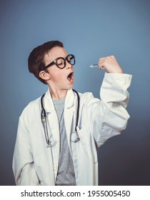 Cool Young Boy Is Disguised As A Doctor With A White Doctor Smock And Plays With Syringe And Stethoscope In Front Of Blue Background