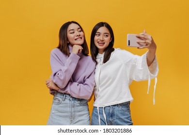 Cool Young Brunette Asian Women In Stylish Sweatshirts Take Selfie, Smile Sincerely And Pose In Good Mood On Orange Background.