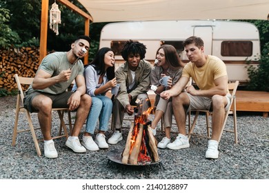 Cool young diverse friends having outdoor picnic near RV, frying marshmallows on campfire, having fun during summer vacation, enjoying camping dessert. Outdoor activities concept - Powered by Shutterstock