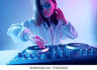 Cool young girl DJ mixes music on a mixing console and headphones, in stylish clothes, glasses on a neon background.