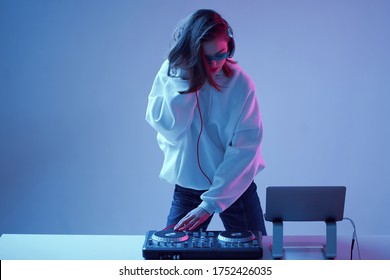 Cool Young Girl DJ Mixes Music On A Mixing Console And Laptop, In Stylish Clothes, Glasses On A Neon Background.