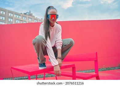 Cool Young Girl Wearing Stylish Modern Outfit, Sporty Sunglasses Posing Squat Sitting On Table Outdoors. Rap Urban Style