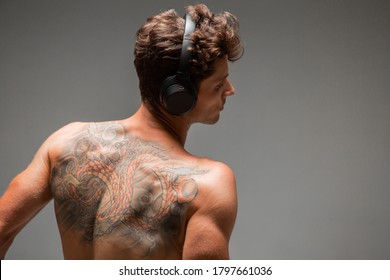 Cool Young Guy In Wireless Headphone Listen To Song With Naked Back Isolated On Gray Background. Body With Tattoo