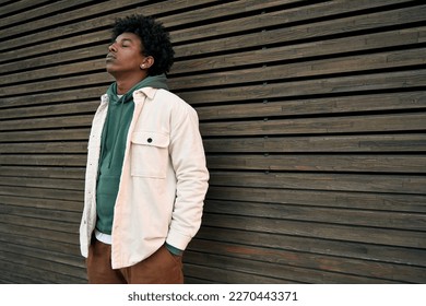 Cool young hipster gen z African American teen standing at wooden wall. Sensitive vulnerable ethnic rebel teenage boy breathing air with eyes closed feeling free or sad in city outdoors. - Powered by Shutterstock