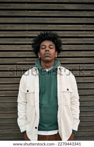Cool young hipster gen z African American teen standing at wooden wall. Sensitive vulnerable ethnic rebel teenage boy breathing deeply with eyes closed feeling stressed or sad in city. Vertical
