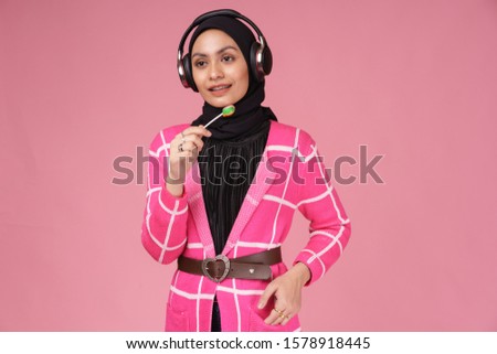 Cool young hipster girl wearing hijab, holding a sucking candy lolipop and listening to music with wireless headphones isolated over pink background. Technology in a modern lifestyle concept.