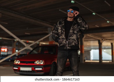 Cool Young Hipster Man With Black Hat In Trendy Clothes With Fashion Military Jacket Straightens His Sunglasses Near A Red Car On The Street