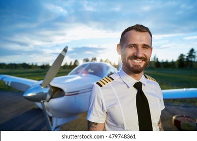 Cool Young Pilot With Tattoos Posing At Small Private Motor Airplane Standing On Runway At Bright Sunset. Blue Sky, Bright Sun