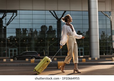 Cool young woman in eyeglasses, beige pants and white blouse walks with yellow suitcase. Charming blonde girl moves near airport with luggage. - Powered by Shutterstock