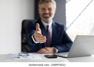 Cooperation Concept. Smiling middle aged businessman extending hand for handshake at camera while sitting at desk in modern office, male entrepreneur in suit offering partnership, greeting companion