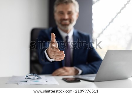 Cooperation Concept. Smiling middle aged businessman extending hand for handshake at camera while sitting at desk in modern office, male entrepreneur in suit offering partnership, greeting companion