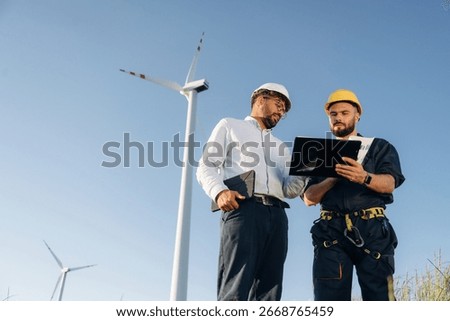 Cooperation concept. Two men engineers are on field, with wind turbines.