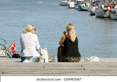 COPENHAGEN, DENMARK - AUGUST 19, 2019: Two Blonde Women,  On The Pier, Near The Water, Enjoying A Relaxing Summer Day In The Copenhagen. Thermos With Coffee. White Cute Dog. Concept Of Hygge.
