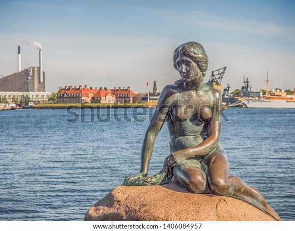 Copenhagen, Denmark\
- May 31, 2018: Bronze statue of the Little Mermaid, symbol of \
Kopenhagen, Den lille Havfrue, on the coastal rocks, with nuclear\
power plant in the\
background