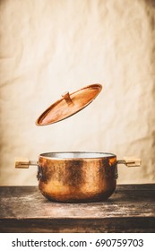 Copper cooking pot with flying  open lid  on wooden kitchen table at wall background, front view