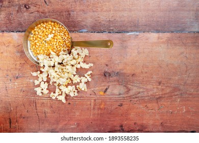 A copper frying pan with unpopped corn kernals in with cooked popcorn overflowing on to a wooden background shot from above with space for text, copyspace, for National Popcorn Day 19 January 2021