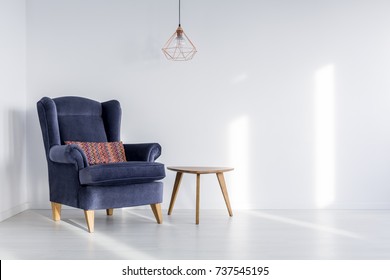 Copper Lampshade Hanging Above Dark Blue Armchair With Colorful Pillow In Room With White Empty Wall