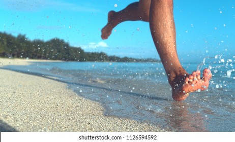 COPY SPACE, LOW ANGLE, DOF: Unrecognizable male tourist jogs on the empty tropical beach and splashes seawater. Active young man on holiday runs barefoot in the wet sand on the coast of exotic island.