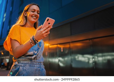 Copy space. One happy young girl using her smartphone to browse the internet while walking on the street at night. Female watching the social media with her cellphone. Girl typing on a mobile phone