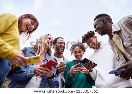 Copy space photo of group of international Erasmus colleagues, standing outdoors connected to their phones, laughing and chatting together. Millennial generation. Low angle view.