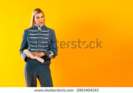 Copy space shot of blonde on yellow background holds a book. Blonde hair model girl posing in old-style military uniform while holding a book. Advertising, education and literature concept. 
