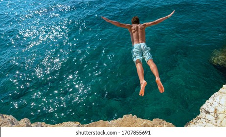 COPY SPACE: Young man on relaxing summer vacation does cliff diving on a hot and sunny day. Cinematic shot of an athletic Caucasian man jumping off a rocky ledge and into the glistening blue ocean.