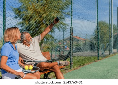 copyspace senior tennis couple sits on a bench by the court, smiling as they take a selfie with a smartphone during a game break. Their relaxed expressions capture a joyful moment of camaraderie - Powered by Shutterstock