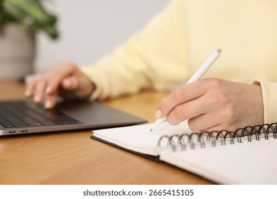 Copywriter using laptop and writing in notebook at wooden table indoors, closeup