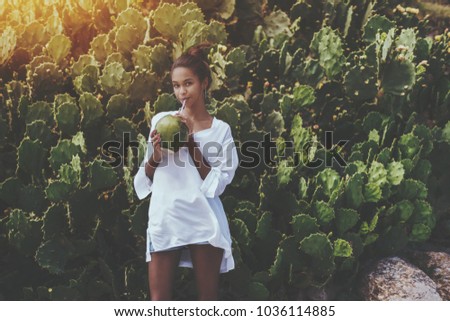 Coquettish young cute Brazilian female in front of quickset hedge of cactuses with coconut is quenching her thirst; black flirtatious girl is drinking coco water while standing near wall of cactuses