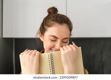 Coquettish young woman smiling, hiding face behind notebook, laughing and smiling, standing in the kitchen.