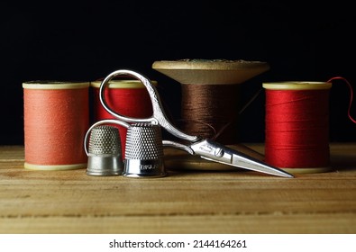 CORAL HUED AND RED COTTON REELS WITH A PAIR OF DECORATIVE SCISSORS AND THIMBLES AND AN OLD VINTAGE WOODEN SPOOL AND THREAD IN BACKGROUND
