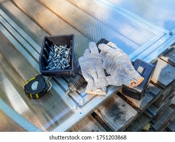 A cordless drill, a tape measure, work gloves, a box of fasteners against the background of cellular polycarbonate for the greenhouse, on wooden pallets. Construction of a greenhouse in the garden.