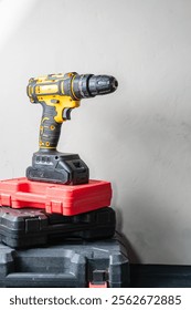 A cordless drill with a yellow and black design, positioned upright on top of red and black toolboxes. The background is a plain gray wall, creating a simple and focused composition.