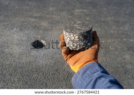 Core sample of road surface material held by a worker in protective glove, with a drilled hole in asphalt. Concept of infrastructure inspection, road repair, and material analysis