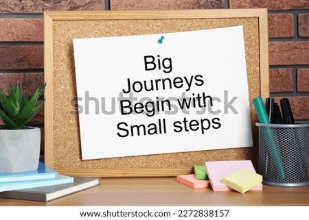 Corkboard and phrase Big Journeys Begin With Small Steps, houseplant, markers and books on table against brick wall. Motivational quote