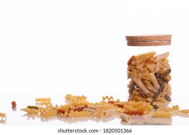 Corked Glass Jar With Uncooked Pasta And Scattered Pasta On Table Surface. Delicious Tasty Organic Whole Grain Italiant Pasta. Italian Mediterranean Cuisine Isolated On White Background Copy Space