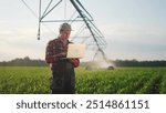 corn agriculture. male farmer works on a laptop in a field with green corn sprouts. corn is watered by irrigation sunlight machine. irrigation agriculture business concept