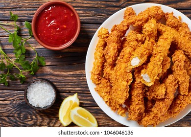 corn flakes breaded deep fried chicken crispy breast strips on white plate, on old rustic  wooden table with tomato sauce and lemon slices, easy recipe for outdoor picnic or party, close-up