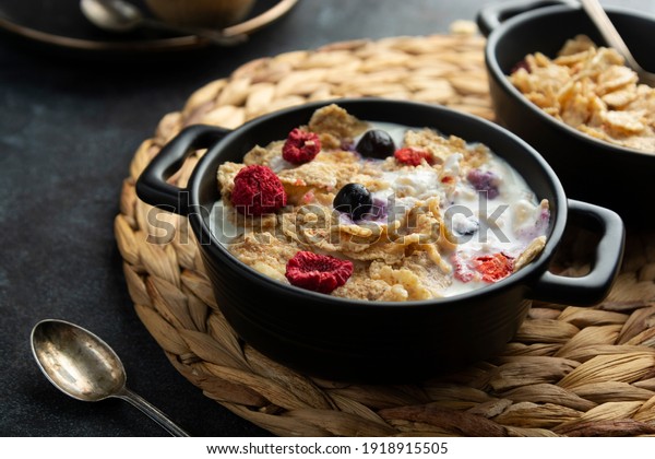 Corn flakes with strawberry and blueberry in\
black bowls over dark\
background