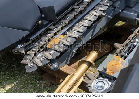 Corn header chain drive with linked feeding plates positioned beside a hydraulic cylinder and lubricated mechanical joints. The assembly includes rotating and structural components used for moving and directing corn plants into agricultural harvesting machinery. Mechanical elements form a rugged component designed for corn harvesting.