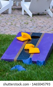 corn hole board with yellow and blue bean bags on purple and navy blue game board outdoors in grass with white lawn chair brick porch background neighborhood party