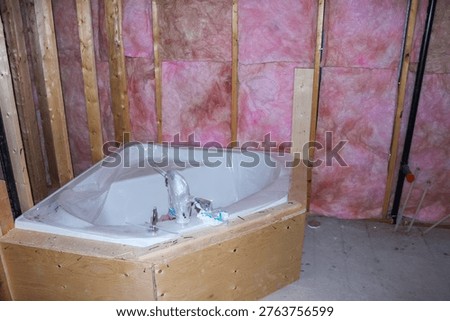 Corner bathtub is positioned in a partially constructed bathroom, featuring exposed wooden framing and pink insulation on the walls