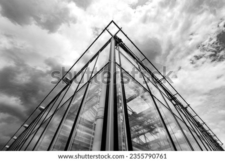 Corner of a glass house with wide angle frog perspective. Vivid cloudscape and sunlight reflected in the windows. Symmetrical architecture in black and white greyscale with high contrast.
